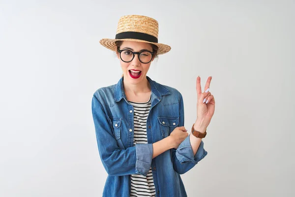Redhead woman wearing denim shirt glasses and hat over isolated white background smiling with happy face winking at the camera doing victory sign. Number two.