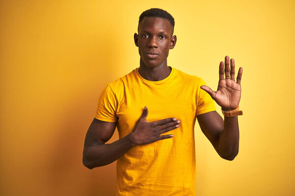 Young african american man wearing casual t-shirt standing over isolated yellow background Swearing with hand on chest and open palm, making a loyalty promise oath