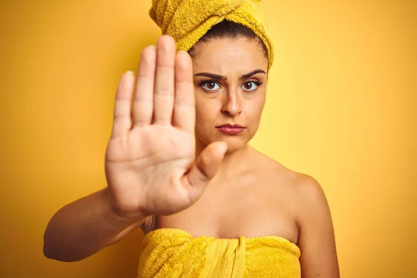 Young beautiful woman wearing towel after shower over isolated yellow background with open hand doing stop sign with serious and confident expression, defense gesture