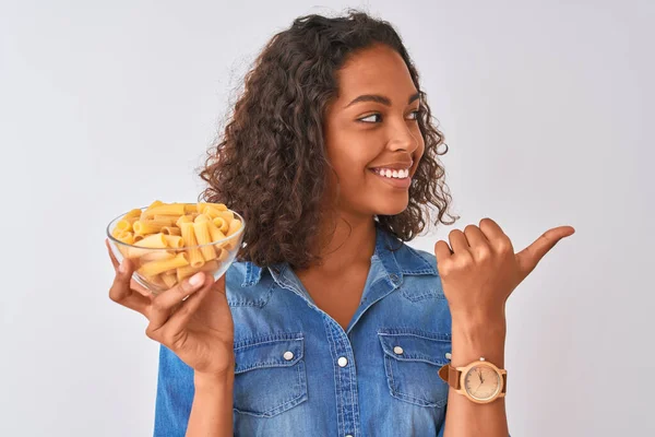 若いですブラジル人女性保持ボウルとともにMacaroniパスタ上の隔離された白い背景ポインティングと表示しますとともに親指で側まで幸せな顔笑顔 — ストック写真