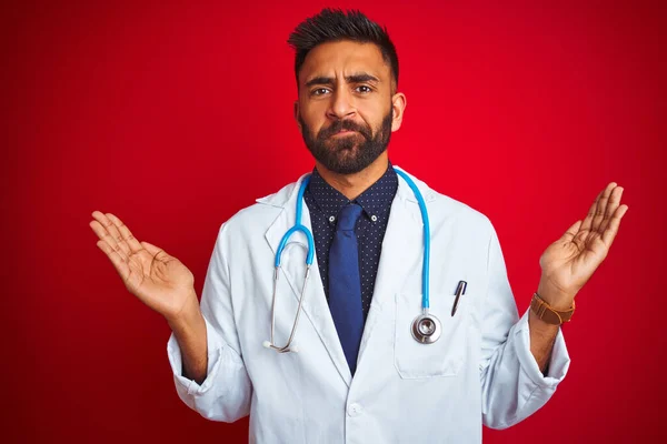 年轻的印度医生 戴着听诊器 站在孤立的红色背景上 毫无头绪 满脸困惑 手臂和双手高举 怀疑的概念 — 图库照片