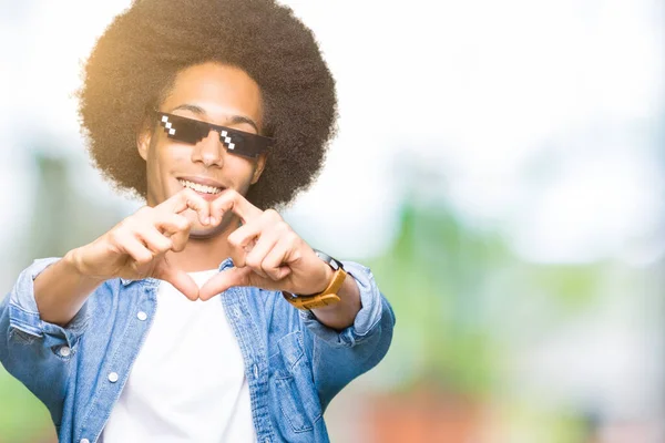Young african american man with afro hair wearing thug life glasses smiling in love showing heart symbol and shape with hands. Romantic concept.