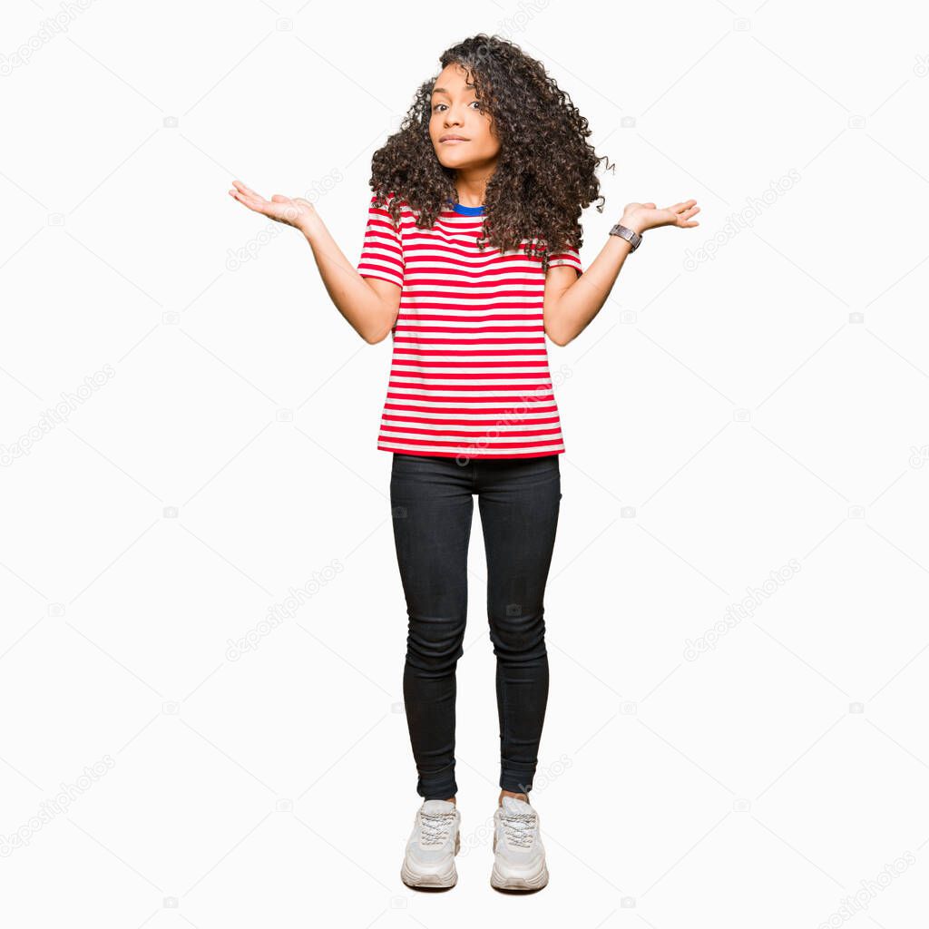 Young beautiful woman with curly hair wearing stripes t-shirt clueless and confused expression with arms and hands raised. Doubt concept.