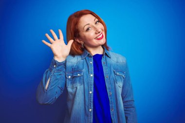 Genç, güzel, kızıl saçlı, kot gömlek giyen, mavi, soyutlanmış arka planda duran, mutlu, güler yüzlü, dostça bir jest yapan bir kadın.