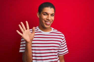 İzole edilmiş kırmızı arka plan üzerinde çizgili tişört giyen genç yakışıklı Arap adam beş numarayı gösterirken kendine güvenen ve mutlu bir şekilde gülümsüyor..