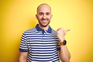 Sakallı genç ve kel bir adam, sarı izole edilmiş arka planda günlük mavi tişört giyiyor, gülümsüyor ve başparmağıyla yana bakıyor..