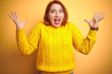 Kışlık kazak giyen güzel bir kızıl kadın izole edilmiş sarı arka planda durmuş çılgın ve başarıyı kollarını kaldırıp heyecanla haykırarak kutluyor. Kazanan konsept