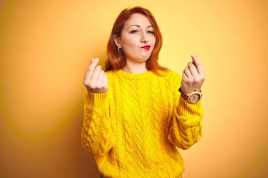 Kışlık kazak giyen güzel bir kadın izole edilmiş sarı arka planda duruyor. Elleriyle para hareketi yapıyor. Maaş ödemesi istiyor.