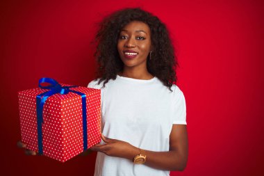 Genç Afrikalı Amerikalı bir kadın doğum günü hediyesi tutuyor. Kırmızı arka planda duruyor. Mutlu bir yüzü var ve gülümsüyor.
