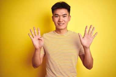 Asyalı genç bir Çinli tişört giyiyor. İzole edilmiş sarı arka plan üzerinde duruyor. Dokuz numarayı işaret ediyor ve gülümsüyor..