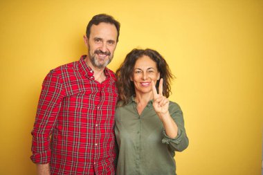 Güzel orta yaşlı çift, izole edilmiş sarı arka planda iki numaralı parmaklarını gösterip gülümserken kendinden emin ve mutlu görünüyorlar..