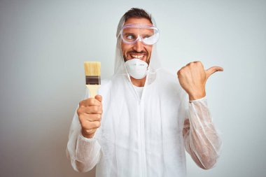 Profesyonel işçi ekipmanı giyen ressam arka planda fırça tutuyor ve gülümseyen bir yüzle baş parmağıyla gösteriyor.