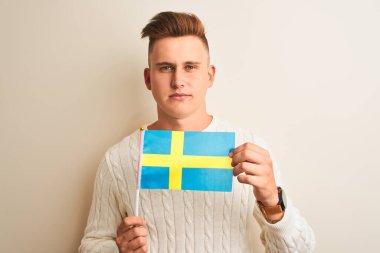 İsveç bayrağını izole edilmiş beyaz arka planda tutan yakışıklı genç bir adam. Zeki bir yüz ifadesiyle ciddi ciddi düşünüyor.