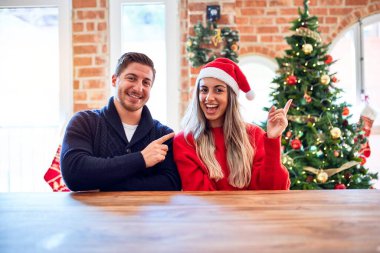 Noel Baba şapkası takan genç bir çift evdeki Noel ağacının etrafındaki sandalyeye ve masaya oturuyor. Yüzlerinde kocaman bir gülümseme, elleri ve parmakları kameraya bakıyor..