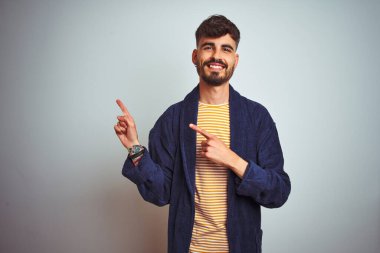 Bornozlu genç adam izole edilmiş beyaz arka plan üzerinde duruyor gülümsüyor ve kameraya bakıyor iki eliyle ve parmaklarıyla kenarı işaret ediyor..