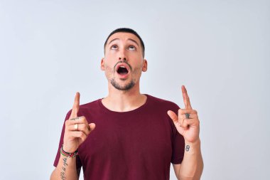 Soyutlanmış arka planda duran yakışıklı genç adam şaşırmış ve şaşırmış bir şekilde yukarı bakıyor ve parmaklarıyla işaret ediyor ve kollarını kaldırıyor..