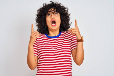 İzole edilmiş beyaz arka plan üzerinde çizgili tişört giyen kıvırcık saçlı genç Arap kadın şaşırmış ve şaşırmış bir şekilde parmaklarıyla işaret ediyor ve kollarını kaldırıyordu..