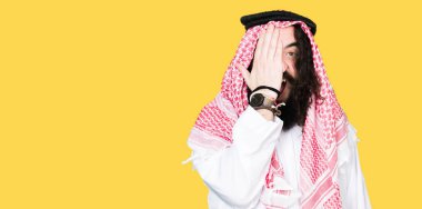 Uzun saçlı Arap iş adamı, yüz ve sürpriz duygu üzerinde emin bir gülümseme ile el ile bir göz kapsayan geleneksel Puşi eşarp giyiyor.