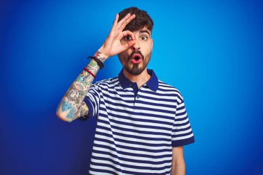 İzole edilmiş mavi arka plan üzerinde duran çizgili polo giyen genç adam şaşırmış yüz ve parmaklarından bakan gözlerle şok olmuş bir jest yapıyor. İnançsız ifade.