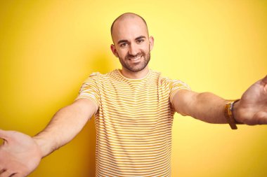 Sarı izole arka plan üzerine sıradan çizgili tişört giyen sakallı genç kel bir adam sarılmak için kollarını açmış gülümseyen kameraya bakıyor. Mutluluğu kucaklayan neşeli bir ifade..