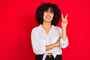 Kıvırcık saçlı, beyaz gömlekli, izole edilmiş kırmızı arka planda gülümseyen, mutlu yüzlü, zafer işareti yapan genç bir Arap kadın. İki numara..