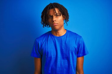 Rastalı tişörtlü Afro-Amerikan bir adam izole edilmiş mavi arka plan üzerinde duruyor şüpheci ve sinirli, sorun yüzünden üzgün. Negatif kişi.