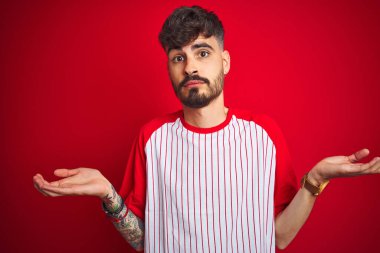 İzole edilmiş kırmızı arka plan üzerinde duran çizgili tişörtlü genç adam elleri ve kolları havada, kafası karışmış bir ifade takınıyor. Şüphe kavramı.