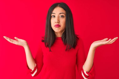 İzole edilmiş kırmızı arka planın üzerinde dikilen sıradan bir elbise giyen genç, güzel Çinli bir kadın. Kolları ve elleri havada, kafası karışmış bir ifade. Şüphe kavramı.