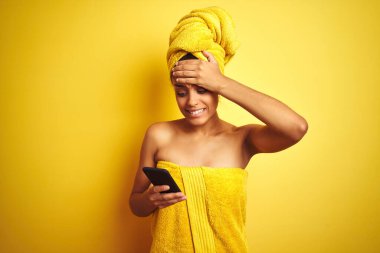 Afro woman wearing towel after shower using smatrphone over isolated yellow background stressed with hand on head, shocked with shame and surprise face, angry and frustrated. Fear and upset for mistake.