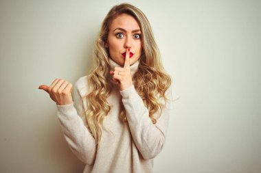 İzole edilmiş beyaz arka planda boğazlı kazak giyen güzel bir kadın sessiz olmak istiyor. Parmaklarını dudaklarına uzatıp elini kenarı işaret ediyor. Sessizlik ve gizli konsept.