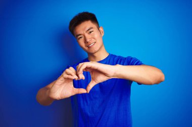 Asyalı genç bir Çinli tişört giyiyor. Mavi arka planda duruyor. Aşkla gülümsüyor, elleriyle kalp sembolü yapıyor. Romantik konsept.