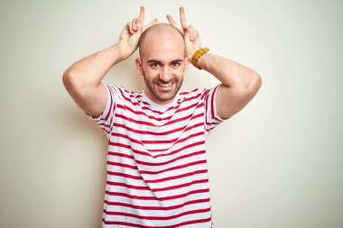 Sakallı, beyaz izole edilmiş arka plan üzerine sıradan kırmızı tişörtlü genç kel bir adam komik ve çılgın pozlar veriyor, parmakları kafasında tavşan kulağı, neşeli bir gülümseme