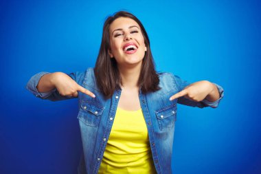 Genç güzel esmer kadın mavi izole arka plan üzerinde rahat mavi kot ceket giyen yüzünde gülümseme ile emin görünümlü, parmakları gururlu ve mutlu kendini işaret.