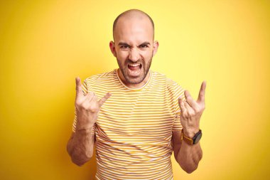 Sarı izole arka plan üzerine günlük çizgili tişört giyen sakallı genç kel bir adam çılgın bir ifadeyle elleri havada rock sembolü yaparak bağırıyor. Müzik yıldızı. Ağır müzik konsepti.