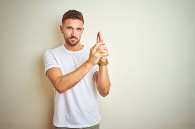 İzole arka planda rahat beyaz t-shirt giyen genç yakışıklı adam el jest ile sembolik silah tutarak, öldürme çekim silah oynarken, öfkeli yüz