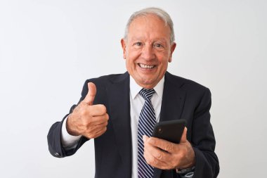 Üst düzey gri saçlı işadamı, izole edilmiş beyaz arka planda duran akıllı telefonu kullanıyor. Büyük bir gülümsemeyle mutlu. Tamam işareti yap, parmaklarınla başparmağını kaldır.