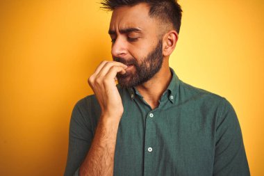 İzole edilmiş sarı arka plan üzerinde duran yeşil tişörtlü genç Hintli adam elleri ağız yiyen tırnaklarla gergin ve stresli görünüyor. Endişe sorunu.