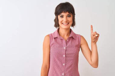 İzole edilmiş beyaz arka planda duran kırmızı tişörtlü genç ve güzel bir kadın bir numaralı parmağını gösterirken kendinden emin ve mutlu bir şekilde gülümsüyor..
