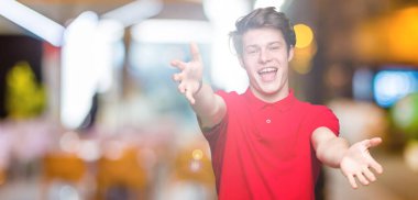 Sarılmak için kollarını açarak gülümseyerek kameraya bakarak izole arka plan üzerinde kırmızı t-shirt giyen genç yakışıklı adam. Neşeli ifade kucaklayan mutluluk.