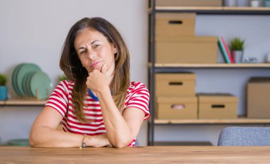 Orta yaşlı bir kadın evinde oturmuş kameraya kendinden emin bir şekilde bakıyor kollarını kavuşturmuş ve çenesini kaldırmış gülümsüyor. Olumlu düşünüyorum..