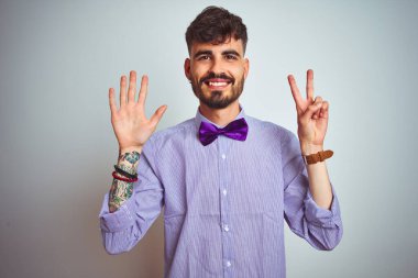 Mor gömlekli ve papyonlu genç adam izole edilmiş beyaz arka planda kendini beğenmiş ve mutlu bir şekilde gülümserken yedinci parmaklarıyla işaret ediyor..
