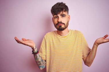 Sarı çizgili tişört giyen genç adam izole edilmiş pembe arka plan üzerinde duruyor. Kolları ve elleri havada, kafası karışmış bir ifade takınıyor. Şüphe kavramı.