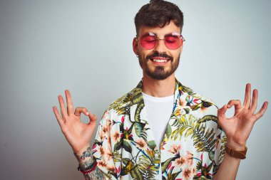 Tatilde dövmeli bir adam güneş gözlüğü takıp izole edilmiş beyaz arka planda rahatlıyor ve parmaklarıyla meditasyon hareketi yaparken gözleri kapalı gülümsüyor. Yoga konsepti.