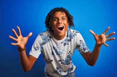 Rasta desenli tişörtlü Afro-Amerikan bir adam izole edilmiş mavi arka plan üzerinde çılgın ve hayret verici bir başarıyı kollarını kaldırarak ve açık gözlerle haykırarak kutluyor. Kazanan konsept