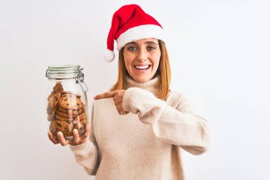 Güzel kızıl saçlı kadın, Noel şapkası takıyor ve arka planda izole edilmiş kurabiye kavanozunu tutuyor. El ve parmağıyla işaret ederek çok mutlu.