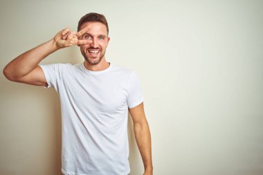 İzole edilmiş arka planda günlük beyaz tişört giyen genç yakışıklı. Yüzünde parmakları olan barış sembolü yapıyor. Neşeli bir şekilde zafere gülümsüyor.