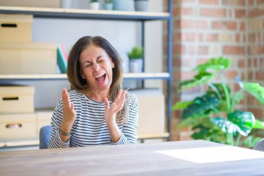 Orta yaş kıdemli kadın evde masada oturan çok mutlu ve heyecanlı silah ile kazanan jest yaparak, gülümseyen ve başarı için çığlık kaldırdı. Kutlama konsepti.