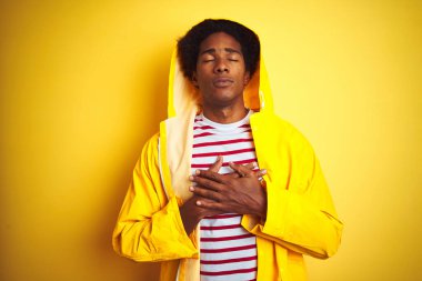 Kapüşonlu bir yağmurluk giyen Afrikalı adam izole edilmiş sarı arka planda duruyor elleri göğsünde, gözleri kapalı ve yüzünde minnettar bir jest ile gülümsüyor. Sağlık kavramı.