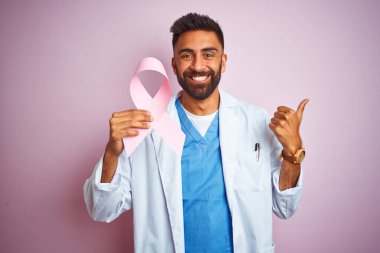 Kanser kurdelesi tutan genç Hintli doktor pembe arka planda duruyor. Kocaman gülümsüyor ve gülümsüyor. İşaret veriyor.