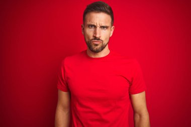 Kırmızı izole edilmiş arka plan şüphecisi ve gergin üzerine gündelik tişört giyen genç yakışıklı bir adam sorun yüzünden asık suratlı. Negatif kişi.
